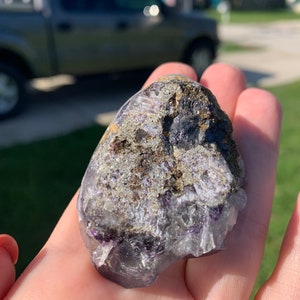 Op de afbeelding: Een hand houdt een ruwe, natuurlijke steen vast. De steen heeft een mix van kleuren, waaronder paars, grijs en bruin. De textuur lijkt een mix te zijn van kristallijne en ruwe oppervlakken. De steen wordt tegen een vage buitenachtergrond gehouden.