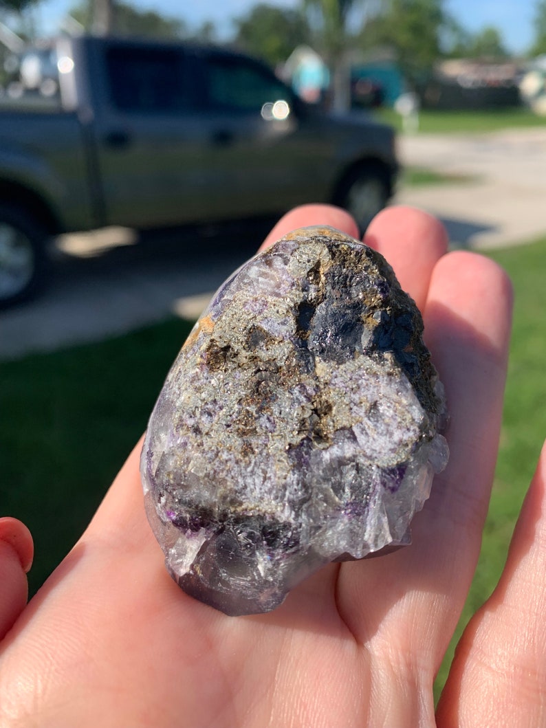 Op de afbeelding: Een hand houdt een ruwe, natuurlijke steen vast. De steen heeft een mix van kleuren, waaronder paars, grijs en bruin. De textuur lijkt een mix te zijn van kristallijne en ruwe oppervlakken. De steen wordt tegen een vage buitenachtergrond gehouden.