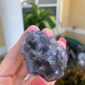 Op de afbeelding: Een close-up van een cluster van fluorietkristallen. De kristallen zijn een mix van donkerpaars en helder, met een ruw, getextureerd oppervlak. De fluoriet wordt vastgehouden in een hand met roze nagellak.