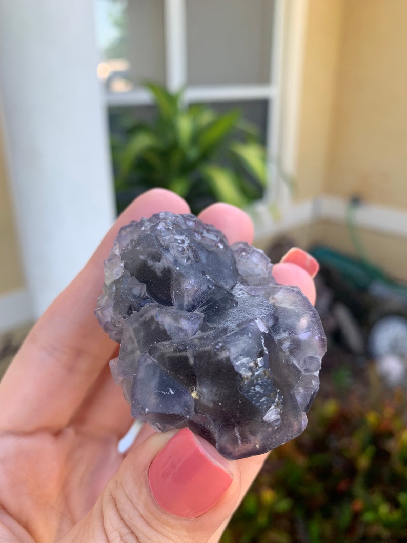 Op de afbeelding: Een close-up van een cluster van fluorietkristallen. De kristallen zijn een mix van donkerpaars en helder, met een ruw, getextureerd oppervlak. De fluoriet wordt vastgehouden in een hand met roze nagellak.