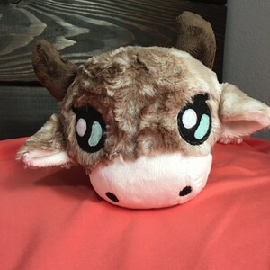 Puede incluir: Una cabeza de vaca de peluche marrón y blanca con grandes ojos azules. La vaca tiene un hocico blanco y cuernos marrones.