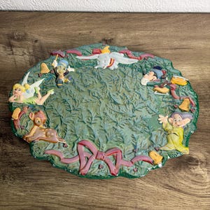 May include: An oval decorative plate with a green leafy design and pink ribbon accents. The plate features colourful, raised figures of Disney characters, including Dumbo, Tinkerbell, and Snow White's dwarfs. The plate is on a wooden surface.