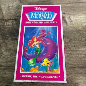 May include: A VHS tape of Disney's "The Little Mermaid: Ariel's Undersea Adventures." The cover features Ariel, Flounder, and a seahorse. The tape has a pink border and the text "Stormy, the Wild Seahorse."