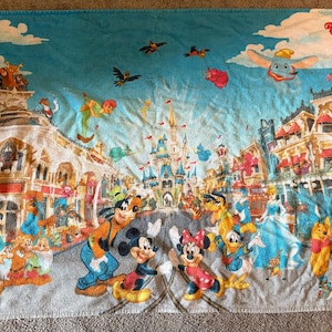 May include: A large, rectangular beach towel featuring a colorful illustration of a theme park scene with cartoon characters. The towel has a blue sky background with the words "The Magic Kingdom" at the top. Mickey and Minnie Mouse are in the center.