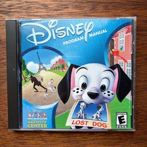 May include: A Disney 102 Dalmatians Activity Center program manual in a clear plastic CD case. The cover features a white puppy with black spots holding a Lost Dog sign in its mouth, with a background showing the Eiffel Tower and a red parrot.