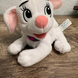 Può includere: Un giocattolo di peluche bianco a forma di cane con orecchie interne rosa, naso rosa e ciglia nere. Il cane ha una bocca rosa e un collare nero e rosa. L'etichetta recita "star bean™" e "MADE IN CHINA © Disney."
