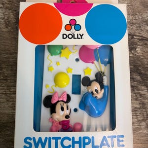 May include: A vintage switchplate featuring Minnie and Mickey Mouse characters. Minnie wears a pink bow and dress, while Mickey is in a blue outfit, holding balloons. The packaging includes the word "SWITCHPLATE" and colorful circles.