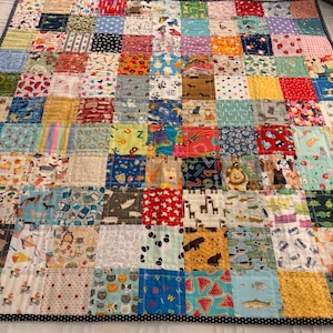 May include: A patchwork quilt featuring a grid of square fabric patches with diverse colorful prints, including animals, fruits, geometric patterns, and letters. The quilt is finished with a thin black and white polka dot fabric border.
