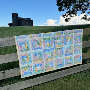 May include: A colorful patchwork quilt with a floral pattern in shades of pink, blue, yellow, and green. The quilt is hanging on a wooden fence against a backdrop of a grassy field and a barn in the distance.