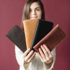 Op de afbeelding: Vier leren portemonnees in verschillende kleuren: bruin, zwart, donkerbruin en beige. De portemonnees worden in de handen van een persoon gehouden.