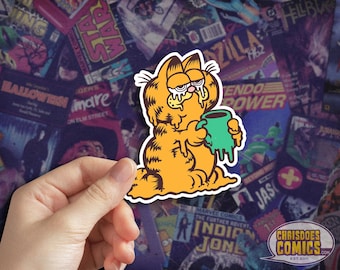 May include: A sticker featuring Garfield, the orange cat, holding a green mug, with a tired expression. The sticker is set against a backdrop of comic book covers. The sticker has a white border and the text "CHRISDOES COMICS" in a speech bubble.