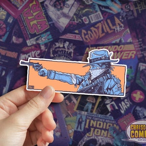 May include: A sticker featuring a cartoon illustration of a person wearing a blue jacket, a bandana, and a cowboy hat, holding a gun. The background is orange.