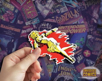 May include: A red and yellow comic book style sticker featuring a woman in a yellow dress lying on the ground with a bloody background. The sticker has a jagged edge and is being held by a hand.