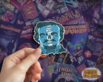 May include: A blue and white sticker featuring a portrait of Edgar Allan Poe with a skull face. The sticker is being held by a hand against a background of comic book covers.