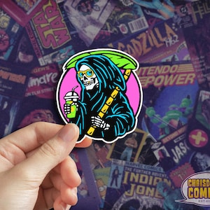 May include: A sticker featuring a cartoon skeleton wearing a black hooded robe and holding a green drink with a straw. The skeleton is holding a yellow and green striped stick with a green leaf on top. The background is pink with a white outline.