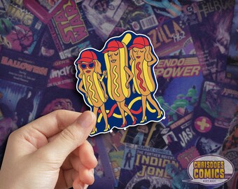 May include: A sticker featuring three cartoon hot dogs dressed as women, wearing red caps and heels. The hot dogs have blonde hair and are outlined in blue. The background is a collage of comic book covers. The sticker is held by a hand.