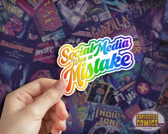 May include: A rainbow-colored sticker with the text "Social Media Was a Mistake" in a bold, stylized font. The sticker is held in a hand against a background of comic book covers.