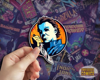 May include: A sticker featuring a stylized illustration of Michael Myers from the Halloween film series. The character is depicted holding a bloody knife, set against an orange and blue background. The sticker is held in a hand, with a comic book background.