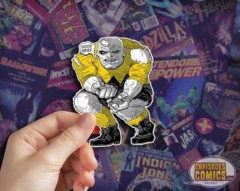 May include: A yellow and black comic book character sticker with the text "Good Grief." The character is a large, green-skinned, humanoid figure with a large head and a menacing expression. The character is in a crouching position with its hands clasped in front of it.