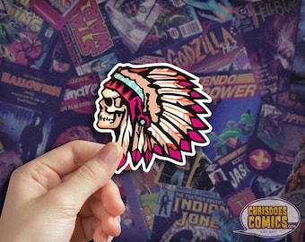 May include: A sticker featuring a skull wearing a Native American headdress in pink, red, and tan. The sticker is held against a backdrop of comic book covers. The sticker has the text "CHRISDOES COMICS EST 2011".