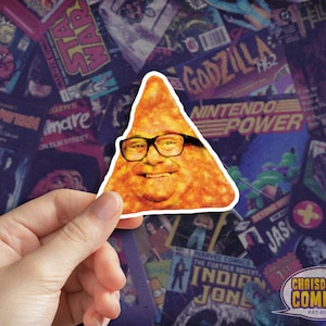 May include: A triangular sticker featuring a face wearing glasses, resembling a Dorito chip. The sticker is held against a backdrop of comic book covers and movie posters. The sticker is from ChrisDoesComics.com.