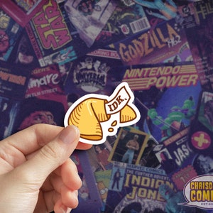 May include: A yellow fortune cookie with the text "IDK" written on it. The cookie is held in a hand against a background of colorful comic book covers.