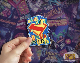 May include: A colorful sticker with the text "Be Kind is Punk Rock" in a Superman-inspired design. The sticker is held against a backdrop of vintage comic book covers, featuring various titles and logos.