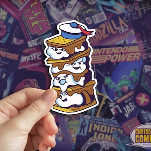 May include: A stack of four s'mores with a sailor hat on top. The s'mores are made with graham crackers, marshmallows, and chocolate. The marshmallows are smiling and have googly eyes. The s'mores are on a white background.