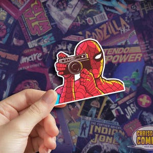 May include: A sticker featuring Spider-Man holding a vintage camera, set against a backdrop of comic book covers. The sticker is red and yellow with a white border. The sticker is held by a hand. The sticker has the text "CHRISDOES COMICS EST 2011".