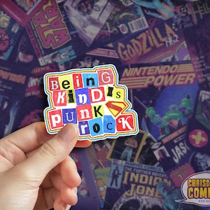 May include: A colorful sticker with the words "Being Kind Punk Rock" in a collage of different fonts and colors. The sticker is held in a hand against a background of vintage comic book covers.