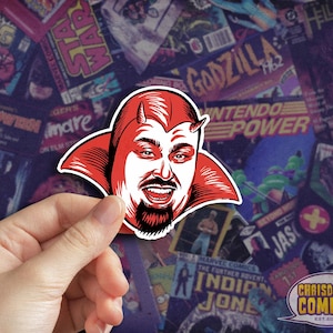 May include: A red and white sticker featuring a smiling devil-like character with horns and a goatee. The sticker is held in front of a background of comic book covers. The sticker is from ChrisDoesComics.