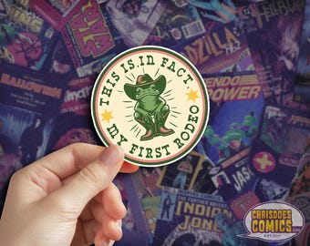 May include: A round sticker with a green frog wearing a cowboy hat and boots. The text "THIS IS, IN FACT, MY FIRST RODEO" is printed around the frog. The sticker has a cream-colored background with a pink and green border.