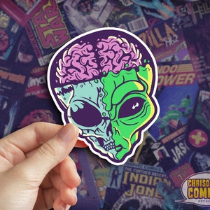 May include: A sticker featuring a green and blue alien with a pink and white brain exposed. The alien has a skull-like face with large eyes and a wide mouth. The sticker is on a background of colorful comic book covers.