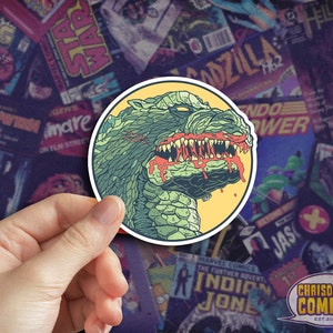 May include: A sticker featuring a green and yellow illustration of a monster with red dripping blood from its mouth. The monster has sharp teeth and a scaly, textured skin. The sticker is circular and has a yellow background.