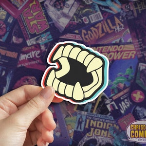 May include: A sticker of a cartoon mouth with sharp teeth, in a 3D style, with red, white, and blue accents. The sticker is being held by a hand against a background of comic book covers.