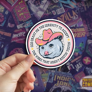 May include: A round sticker featuring a cartoon opossum wearing a pink cowboy hat. The text reads "God grant me the serenity to accept the vibes that aren't rootin' tootin'". The sticker is set against a background of comic book covers.