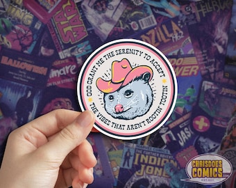 May include: A round sticker featuring a cartoon opossum wearing a pink cowboy hat. The text reads "God grant me the serenity to accept the vibes that aren't rootin' tootin'". The sticker is set against a background of comic book covers.