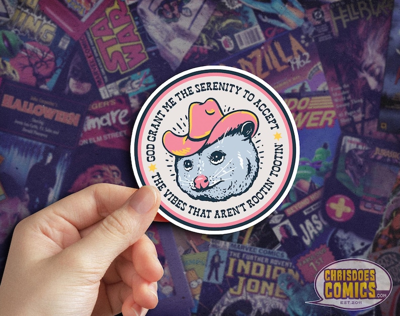 May include: A round sticker featuring a cartoon opossum wearing a pink cowboy hat. The text reads "God grant me the serenity to accept the vibes that aren't rootin' tootin'". The sticker is set against a background of comic book covers.