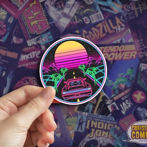 May include: A retro-style sticker featuring a pink and blue sunset, palm trees, and a car driving down a neon-lit road. The sticker is circular and has a black background.