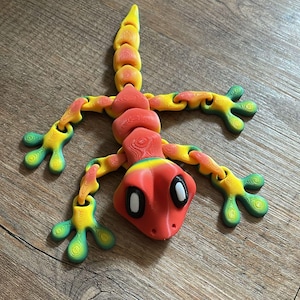 Peut inclure: Un jouet articulé en forme de gecko rouge, jaune et vert. Le gecko a un corps et des membres segmentés, et de grands yeux blancs.