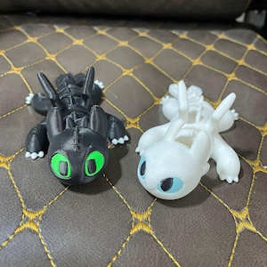 Peut inclure: Deux figurines de dragon imprimées en 3D, l'une noire avec des yeux verts et l'autre blanche avec des yeux bleus. Les dragons sont en position assise, les ailes déployées.