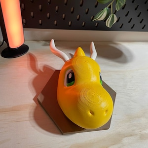 Pode incluir: Uma cabeça de dragão amarela impressa em 3D, montada na parede, com olhos verdes e chifres brancos. A cabeça de dragão está montada em uma base hexagonal marrom.