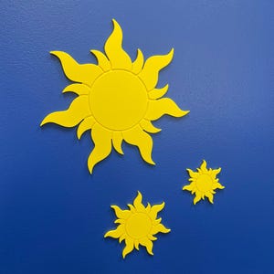 May include: Three bright yellow sun-shaped wall decorations with wavy, flame-like rays extending from a central circular core. The suns are arranged in a cluster, with one large sun at the top left and two smaller suns positioned below and to the right.