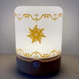 May include: A white glass lamp with a wooden base features a gold sun design with a decorative border. The lamp is illuminated, casting a warm glow.