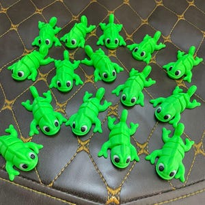 Könnte beinhalten: Eine Ansammlung kleiner, leuchtend grüner Spielzeug-Eidechsen mit schwarz-weißen Augen. Die Eidechsen sind auf einer braunen, strukturierten Oberfläche angeordnet. Jede Eidechse hat einen detaillierten, strukturierten Körper und ausgestreckte Gliedmaßen.
