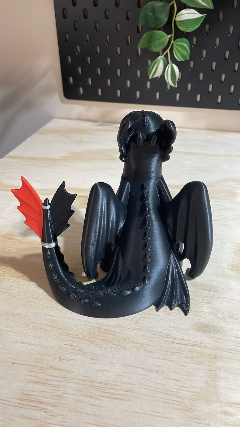 May include: Black 3D printed figurine of a dragon with red wings and a silver tail. The dragon is sitting with its wings spread out.