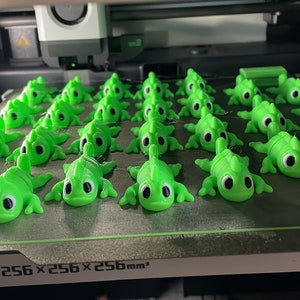 Könnte beinhalten: Ein 3D-Druckbett mit einem Druck von 25 grünen Chamäleon-Figuren. Das Bett ist mit "Build Volume 256 x 256 x 256 mm³" beschriftet.