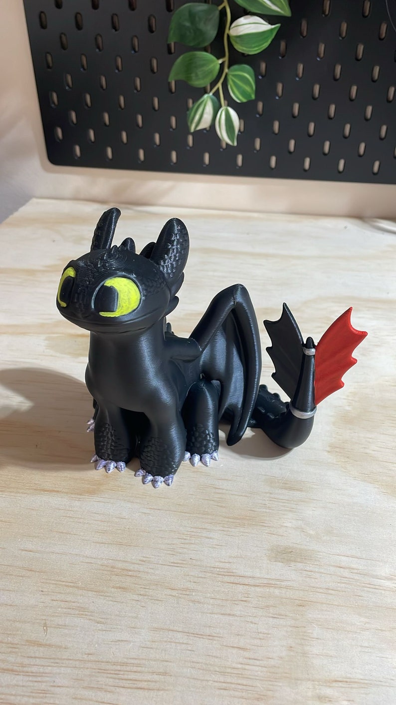 May include: A black 3D printed figurine of Toothless, the Night Fury dragon from the How to Train Your Dragon franchise. The dragon has yellow eyes, a red wing, and a black body with a textured surface.
