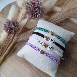 Op de afbeelding: Vier macramé armbanden in groen, zwart, wit en paars, elk met een gouden hartbedel en letterkralen met de letters "KNM". De armbanden worden gepresenteerd op een klein, vierkant, beige kussen.