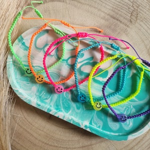 Op de afbeelding: Zeven kleurrijke vriendschapsarmbanden met smiley-bedels. De armbanden zijn gemaakt van gevlochten touw in neon groen, oranje, roze, geel, blauw en paars.